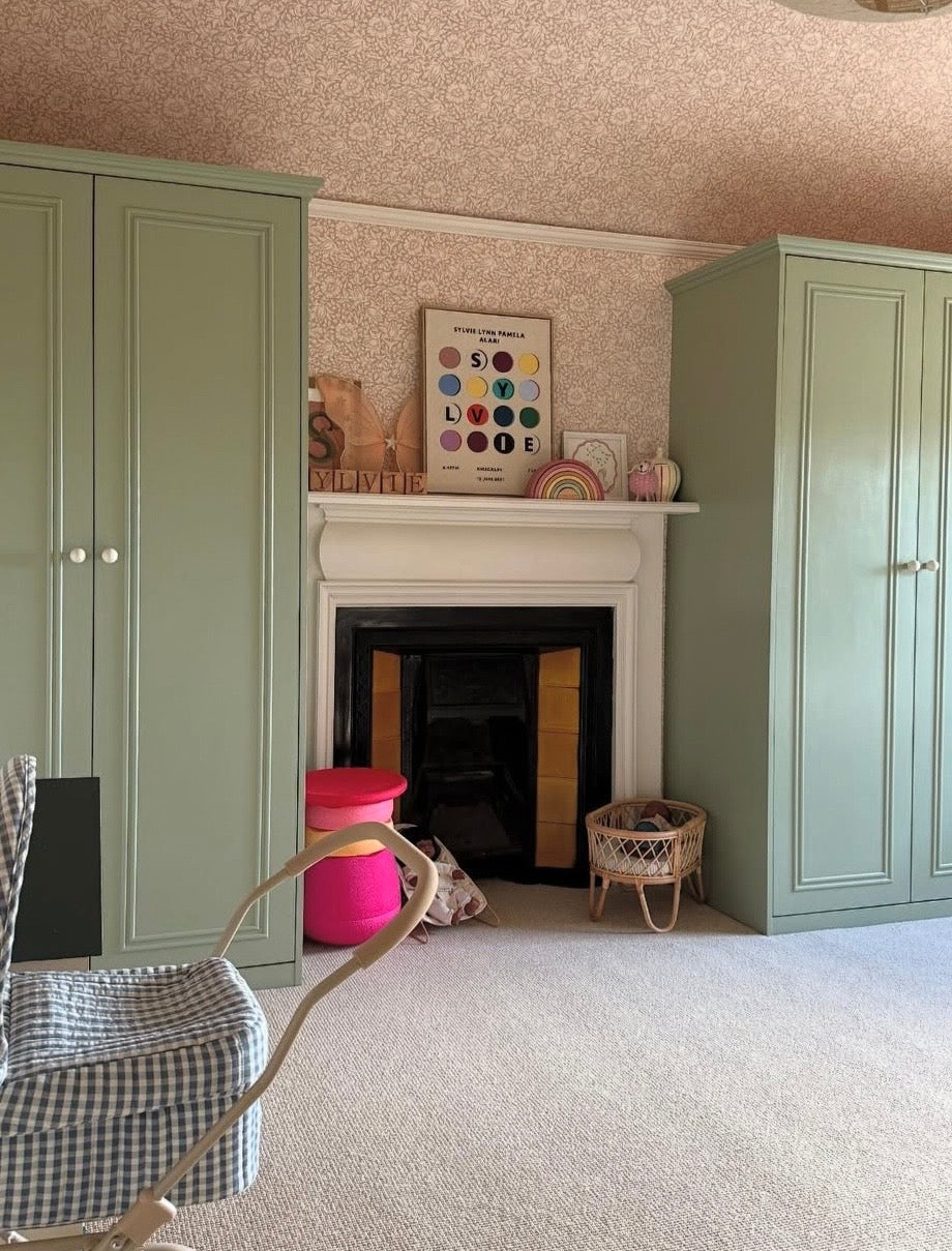 Nursery with pink floral printed wallpaper, artwork above fire place, sage green cupboards