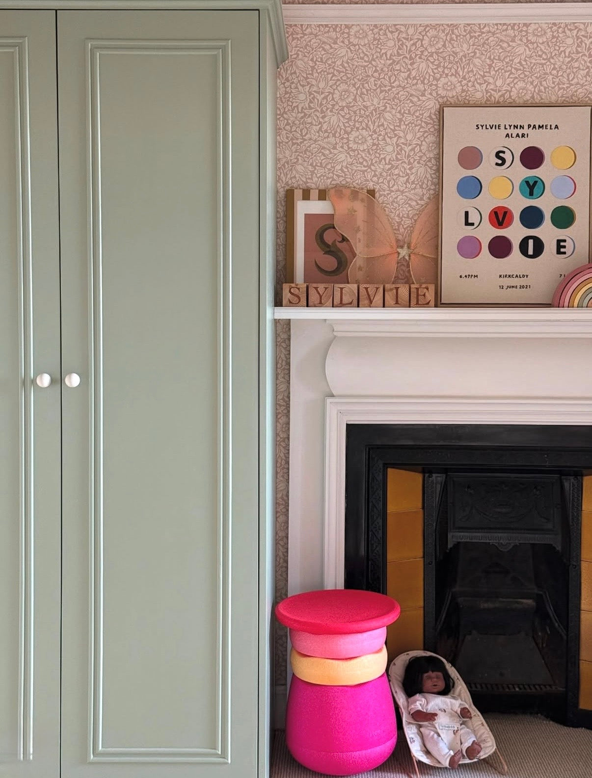 childrens room with pink floral wallpaper, artwork above fireplace, sage green cupboards